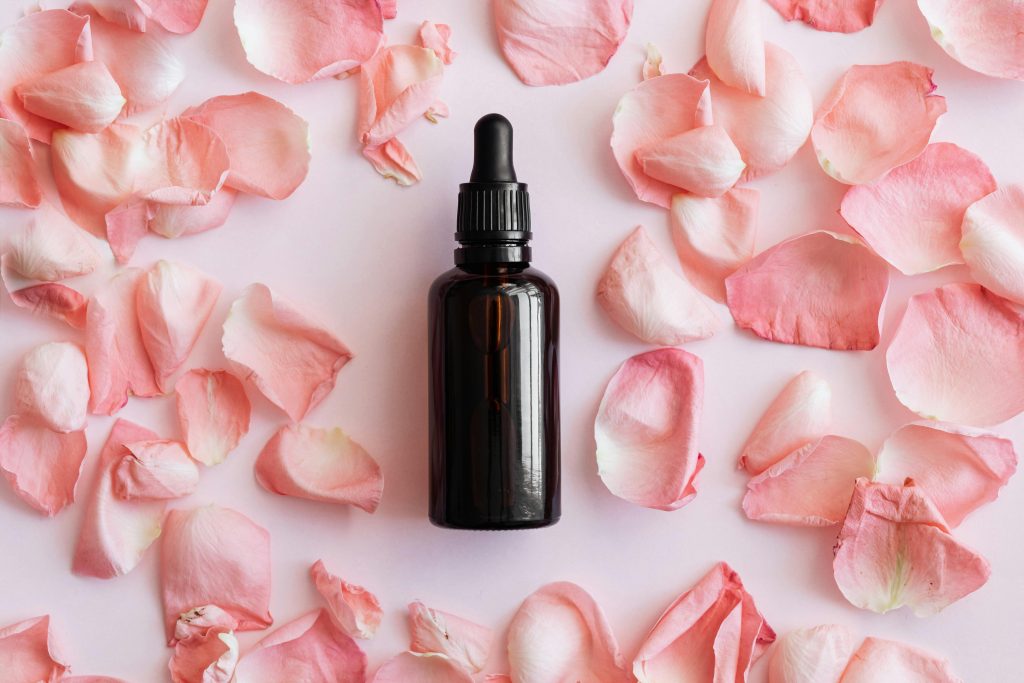 A dark glass dropper bottle surrounded by delicate pink rose petals on a pastel background.